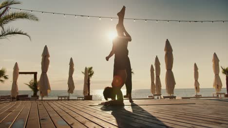 The-guy-helps-his-friend-stand-on-his-head-on-a-special-rug-and-supports-his-legs.-Yoga-classes-and-morning-exercises-together