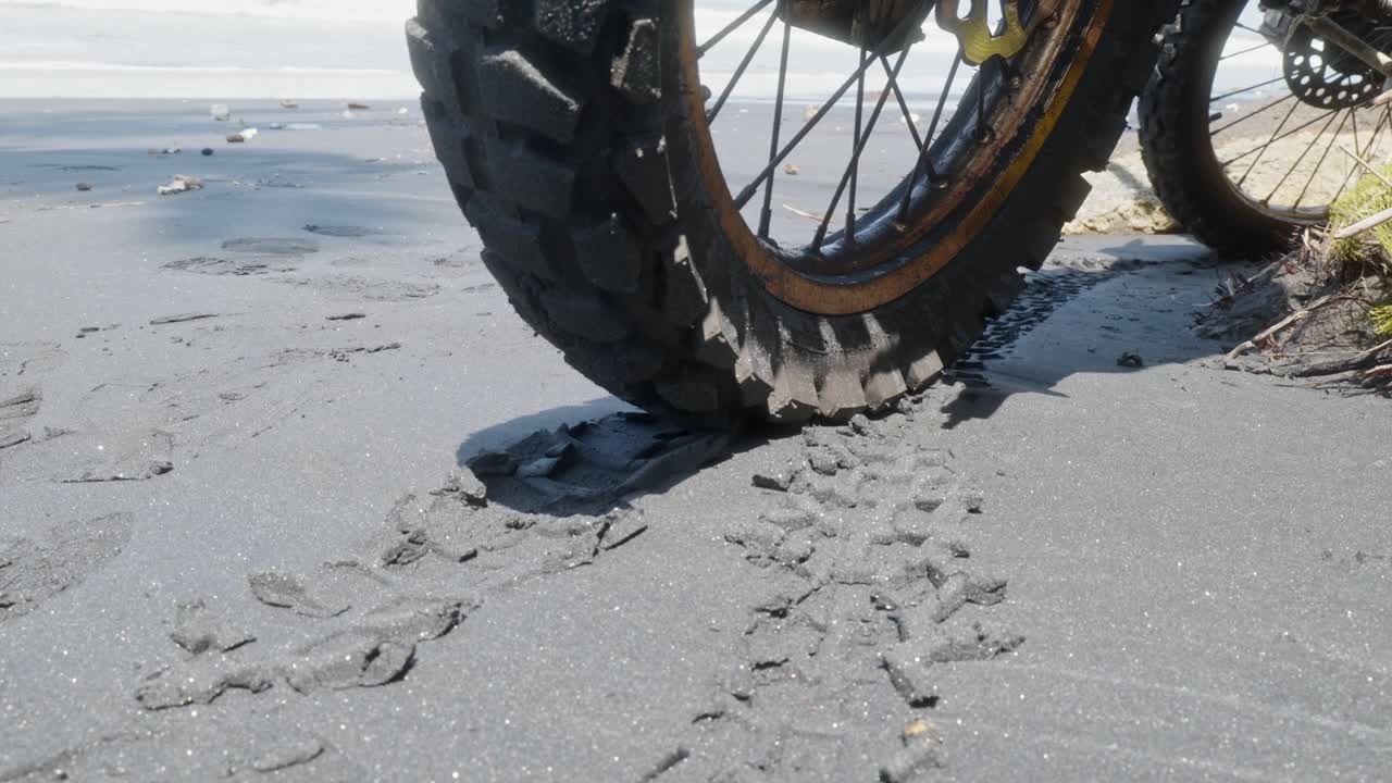 Close-up of a dirt bike tire on black sand, showing fresh tread marks. Shot handheld in bright daylight with beach in background. Gritty and raw perspective