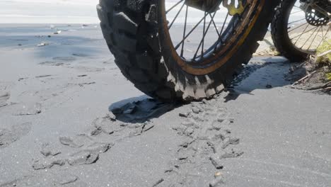 close-up of a dirt bike tire on black sand, showing fresh tread marks. shot handheld in bright daylight with beach in background. gritty and raw perspective