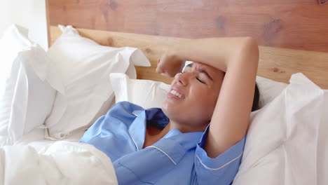 woman in bed wearing blue pajamas using smartphone, looking concerned, at home
