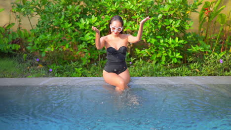 a young women in a one-piece bathing suit sits on the edge of a pool playfully splashing water