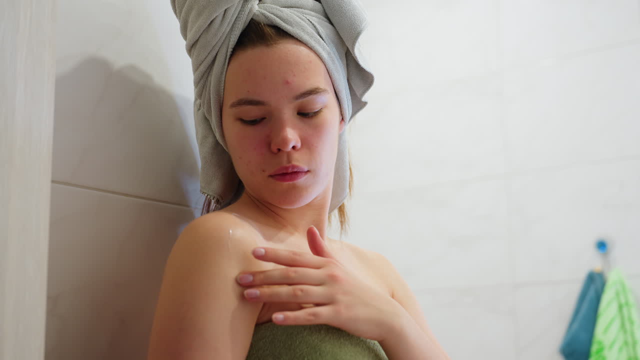 Young girl wrapped in towel applying skincare to her shoulder, part of moisturizing routine, body care, rejuvenation, skin care, healthy skin, smooth skin, self-care, beauty treatment