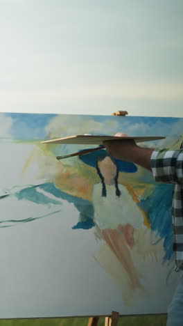un pintor, parcialmente visible, está creando un retrato de una mujer que se mantiene en equilibrio en un vestido blanco y sombrero negro. el escenario es un tranquilo paisaje de hierba