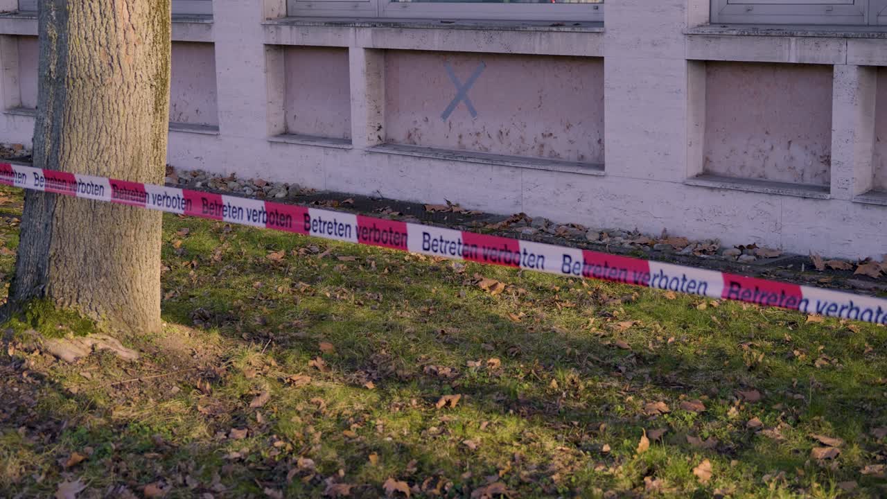 Un ruban rouge et blanc "Interdit d'entrer" entoure un arbre, avec un bâtiment et une fenêtre marquée en arrière-plan.