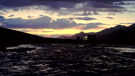 Incline-Hacia-Arriba-Una-Toma-Oscura-Desde-Un-Río-Hasta-Una-Increíble-Puesta-De-Sol-En-Las-Montañas-Con-Siluetas-De-Tiendas-De-Campaña,-Colores-Naranja-Y-Azul-Pintados-En-El-Cielo