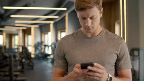 Handsome-sportsman-texting-message-in-sport-club.-Bodybuilder-standing-at-gym