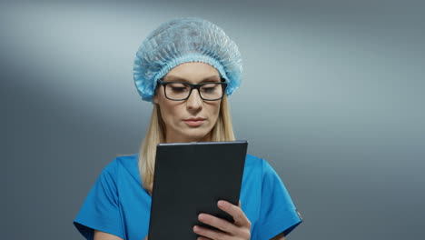 young blonde pretty woman doctor in blue hat and glasses using tablet device, tapping and scrolling on it