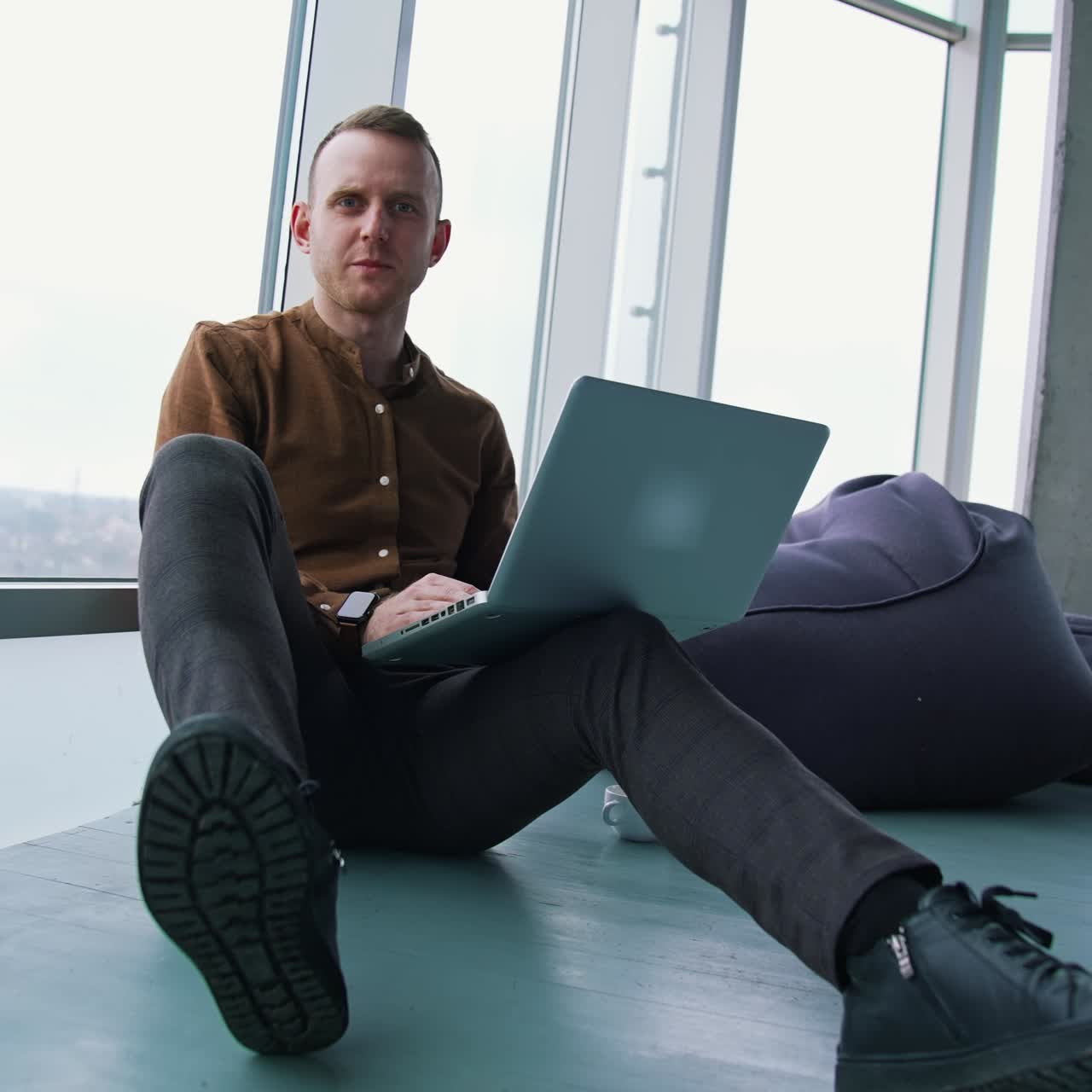 Relaxed businessman in modern office. Young man is sitting on a floor and working on a laptop on windows background. Balanced work