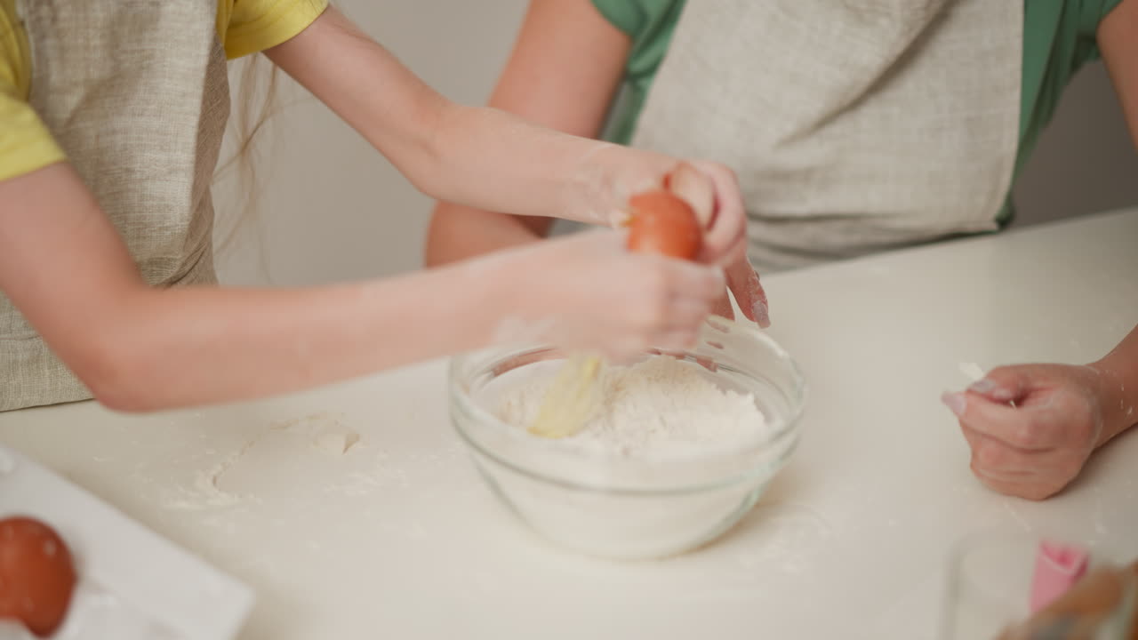 partial view of child holding brown egg over bowl filled with flour while preparing to crack it, flour spread on white counter surface, apron-wearing adult closely supervising hands-on kitchen baking moment