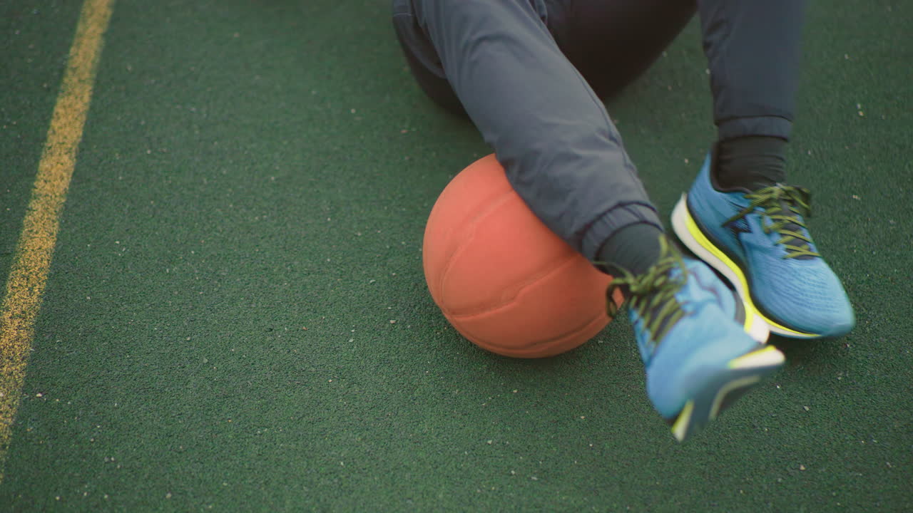 tênis azuis em quadra de basquete verde, close-up da perna de um atleta prendendo uma bola de basquete laranja perto da linha lateral, momento de treino casual com superfície de borracha texturizada, cadarços e sola visíveis, movimento sutil