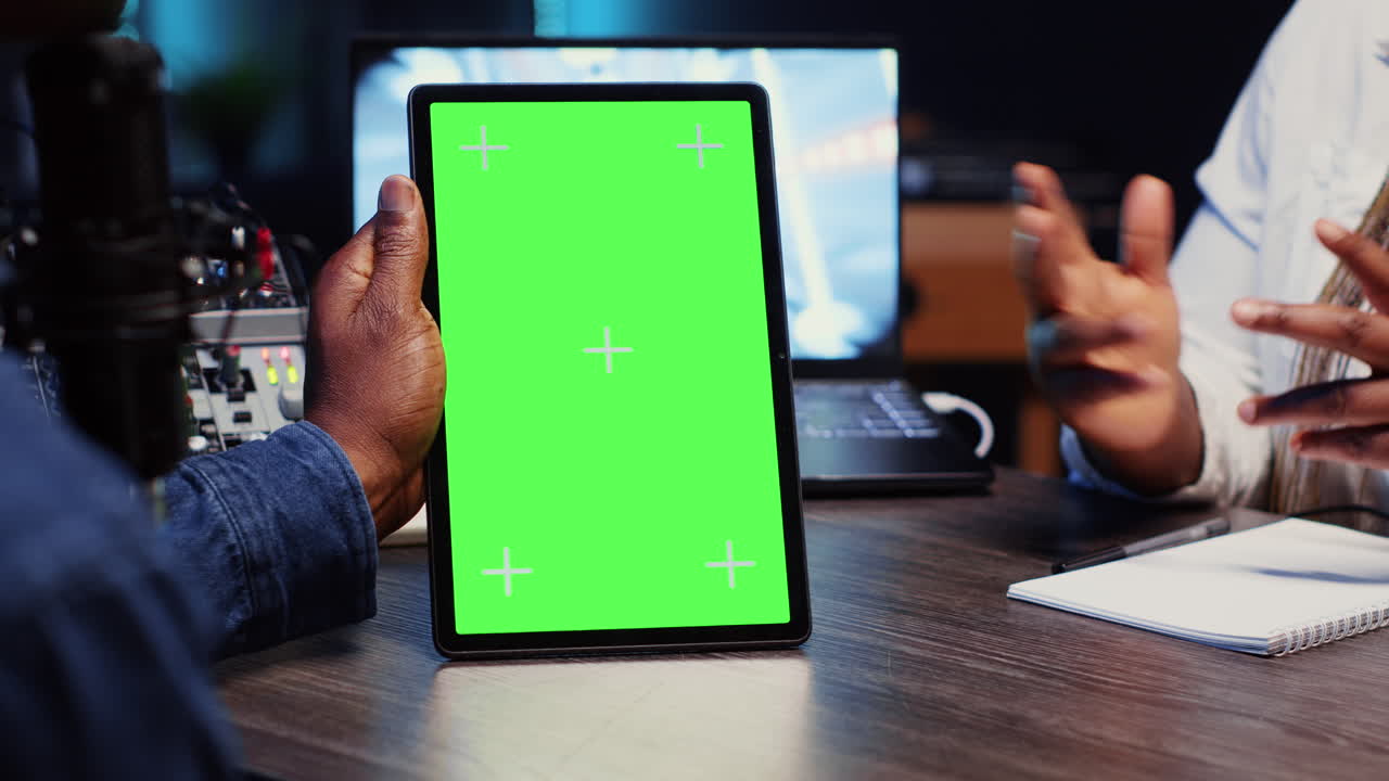 녹색 스크린 태블릿을 들고 있는 블로거 쇼 호스트가 네온 불빛으로 장식된 스튜디오에서 손님과 채팅하고 있습니다.