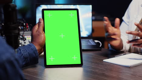 Tableta-De-Pantalla-Verde-Sostenida-Por-El-Presentador-Del-Programa-Vlogger-Charlando-Con-Un-Invitado-En-Un-Estudio-Adornado-Con-Luces-De-Neón