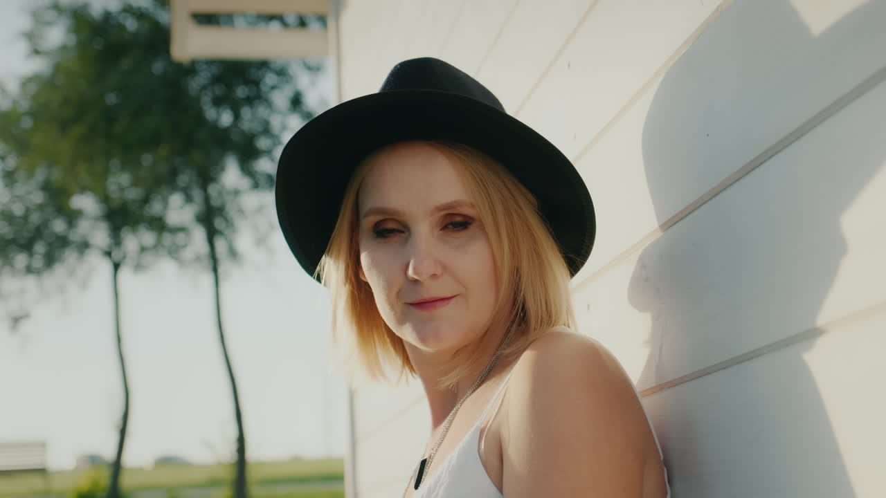 портрет стильной женщины в темной шляпе у белой стены деревянного дома