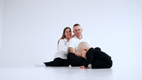 Happy parents look lovingly on the adorable baby sitting on the floor. Little kid is focused on the floor and touches it with his finger.