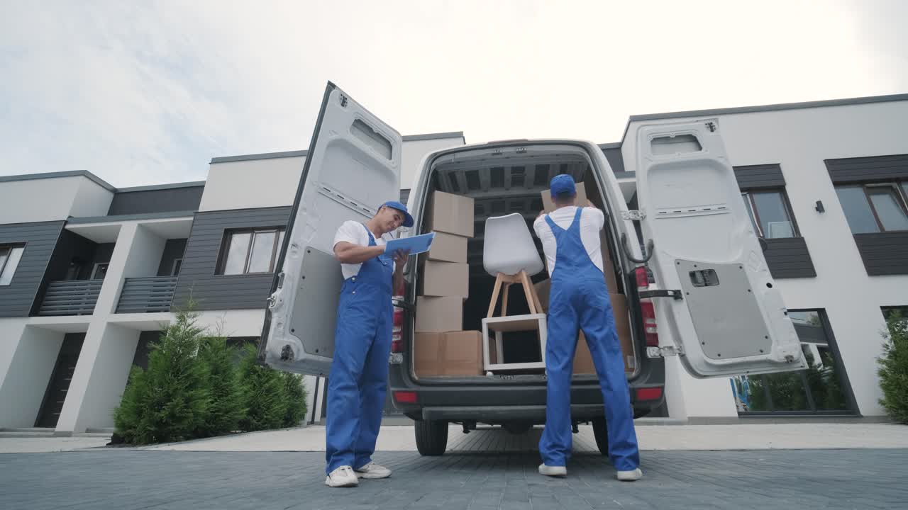 Two young workers of removal company are loading boxes and furniture into a minibus