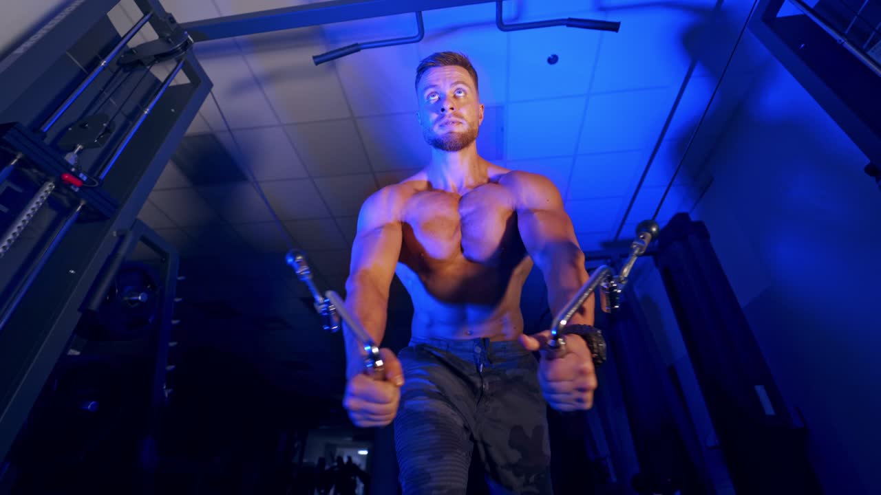 Handsome athlete pumps up his muscles. Naked torso of a sportsman. Muscular bodybuilder training in the gym. View from below. Blue light.