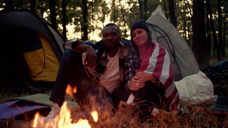 a man with black skin and a girl with a bob in a black hat sit near a fire during a camping stop and hug under the us flag against the backdrop of a beautiful forest and tents