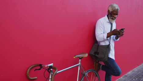 Afroamerikanischer-älterer-Mann-Mit-Fahrrad-Und-Smartphone,-Während-Er-Vor-Einer-Rosa-Wand-Steht