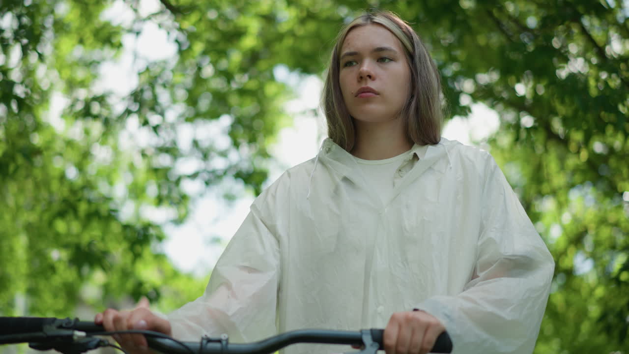 mujer en impermeable translúcido se encuentra al lado de su bicicleta, quitando la mano del manillar mientras toma una respiración profunda, mirando pensativo, rodeado de árboles verdes exuberantes, la luz del sol se filtra a través