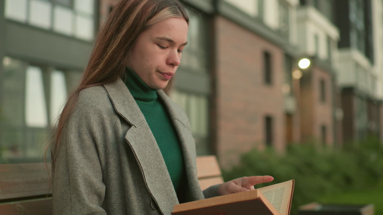 Lady seated on wooden bench outdoors adjusting hair while holding book in hand, blur view of people in distant background near residential buildings with calm and natural urban setting