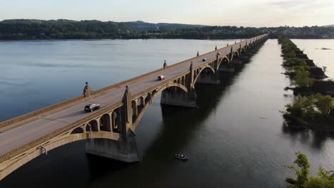 aerial tracking traffic drone shot of susquehanna river bridge at sunset fishing boat on water, former bridge stone piers covered with vegetation and trees