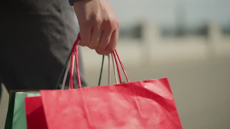 primer plano de un individuo con ropa gris sosteniendo bolsas de compras coloridas mientras camina, la mano agarra bolsas rojas y verdes, con un fondo borroso que da enfoque a la acción de llevar las bolsas