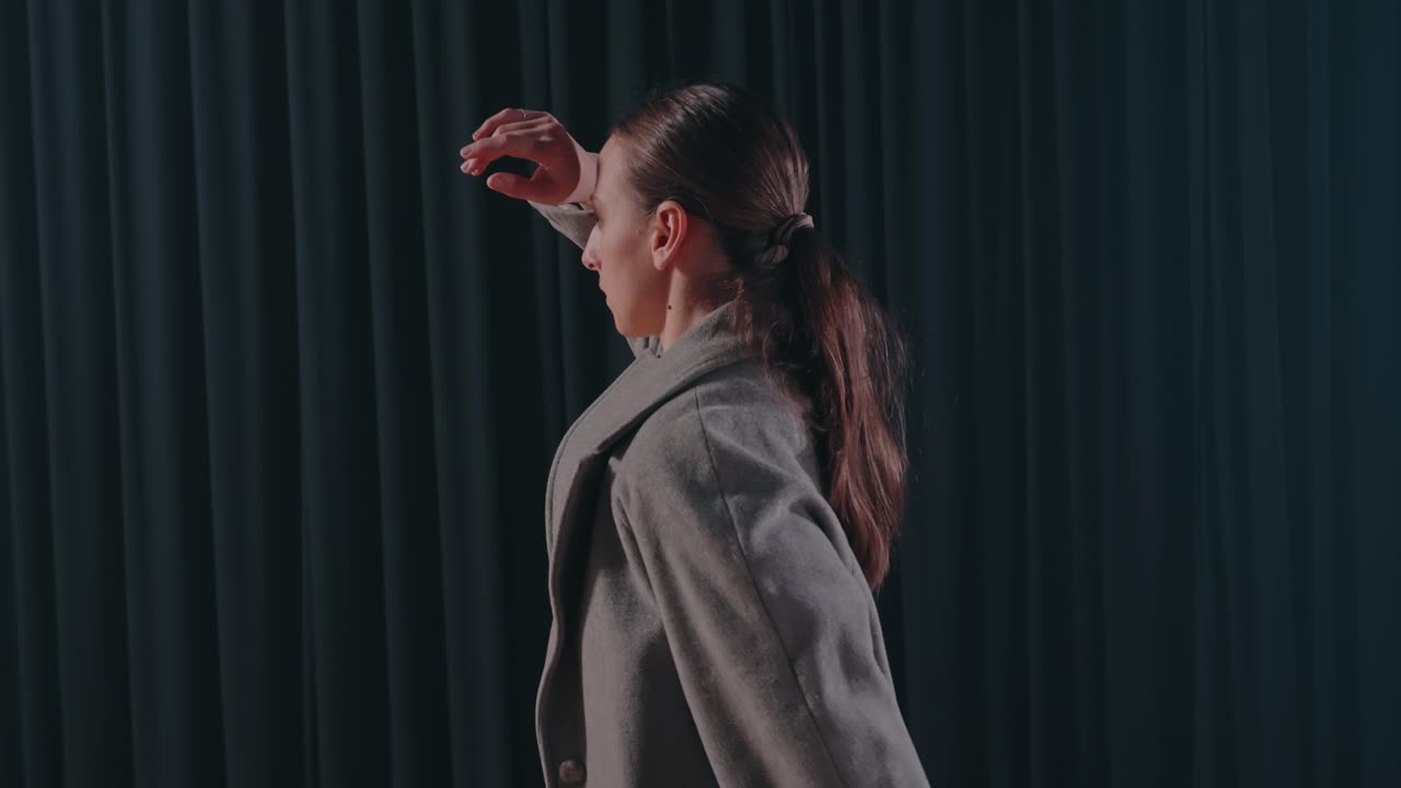 Woman in grey coat performs modern dance routine in front of dark curtain background