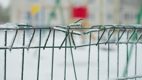 close-up of a green metal fence covered in ice with icicles hanging from the wires, set against a blurred snowy background and urban winter scenery