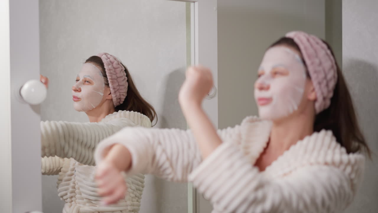 여성은 얼굴 피부를 돌보고 댄스 동작을 수행합니다. 여성은 직물 보습 마스크를 얼굴에 착용하고 휴가를 즐기고 음악을 듣고 느린 동작을합니다.