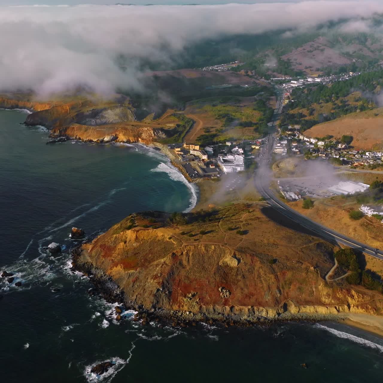 Sunny rocky coastline with houses and highway on top. Thick fog crawling to the city. Aerial perspective