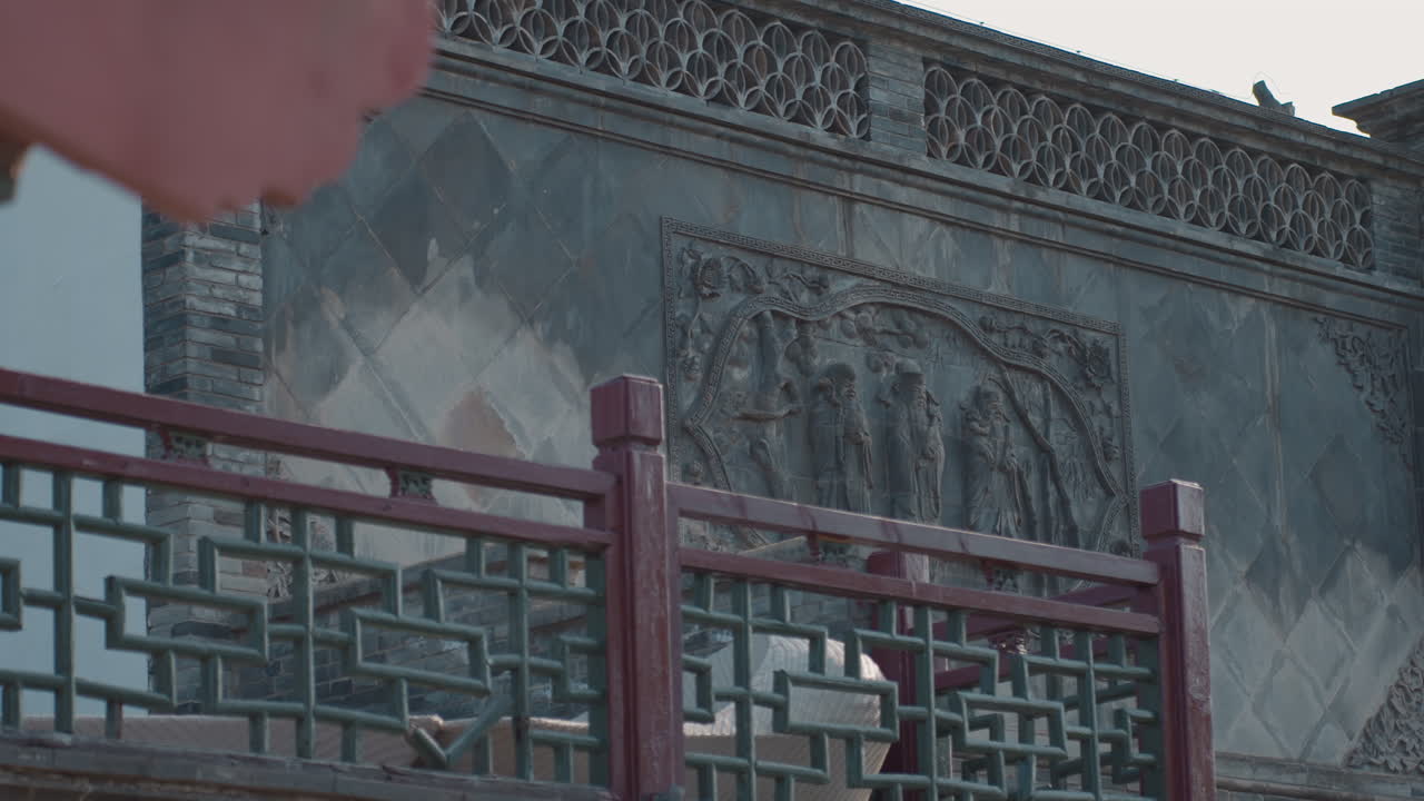 Primo piano di un muro invecchiato di un tempio cinese, la telecamera si muove su una balaustra rossa e un bassorilievo in pietra scolpita grigio-verde, cielo coperto, toni smorzati, rivelazione cinematografica lenta, atmosfera di tranquilla eredità storica.