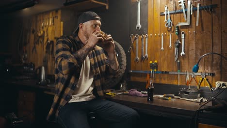 happy-guy-mechanic-in-plaid-shirt-eats-burger-and-drinks-soda-during-lunch-and-break-at-his-work-in-studio-workshop