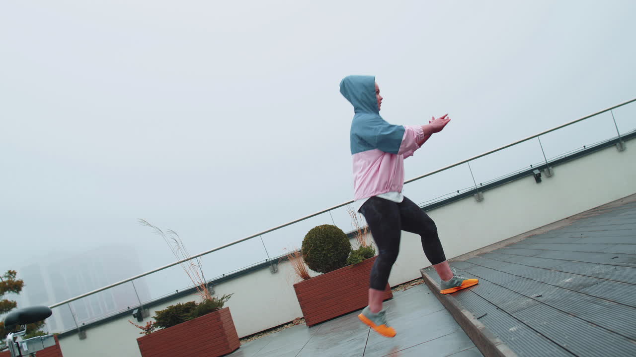 Sport woman in sportswear wears hood doing workout, step cardio exercising training on roof of house