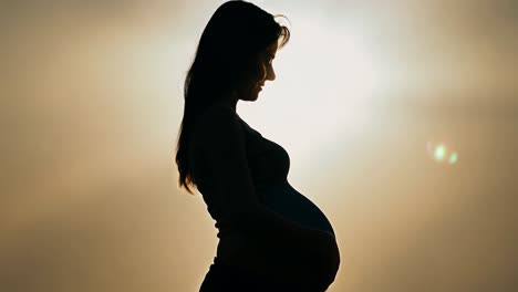 silhouette of a pregnant woman gently touching her belly with her hands against a sunset background