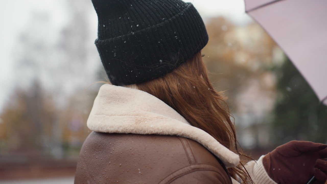 Close view of lady smiling, holding umbrella, wearing knit cap, brown shearling jacket, happily spinning umbrella in light snowfall, surrounded by trees, enjoying autumn day outdoors, joyful expression