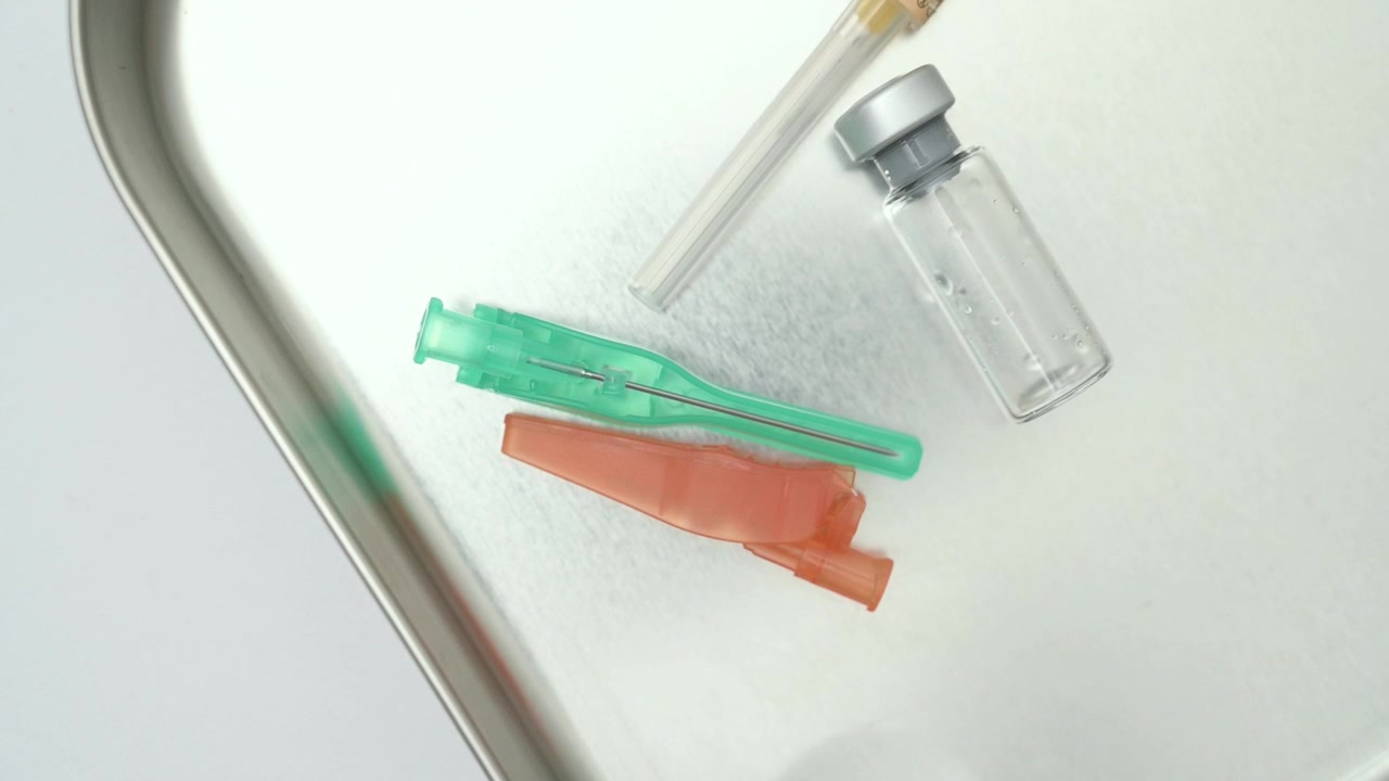 Nurse's gloved hands placing sterile needles and a vaccine vial on a metal tray