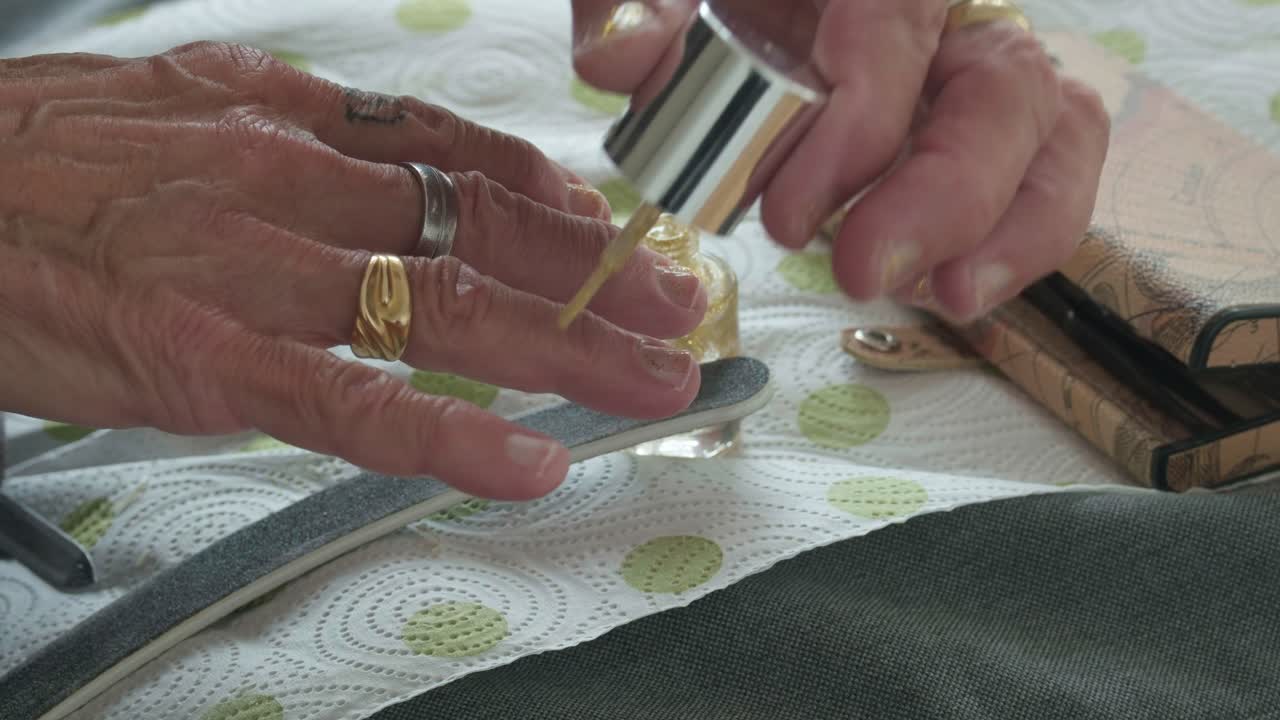 Close-up of senior hands applying shimmering gold nail polish during a home manicure session. Authentic beauty routine featuring jewelry, a nail file, and sparkling gold lacquer detail