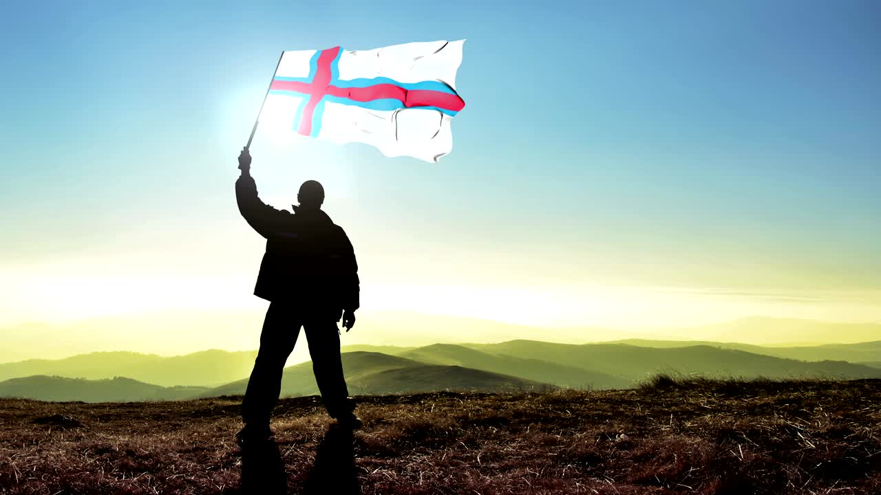 exitoso hombre silueta ganador agitando la bandera de las islas feroe en la parte superior del pico de la montaña. fondo de bucle cinemagraph