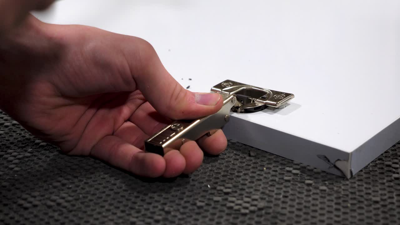 Close-up shot of a hand using a hammer to install a metal hinge on a white wooden panel, set on a textured workbench