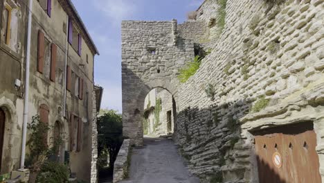 Kleine-Straße-In-Einem-Historischen-Dorf-Mit-Alter-Steinmauer-Und-Torbogen,-Durch-Die-Man-In-Der-Sonne-Frankreichs-Fahren-Kann