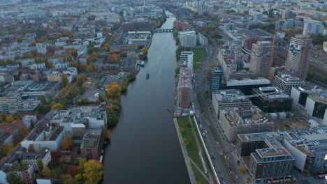 Spree-River-in-Berlin,-Germany-slow-tilt-up-revealing-Cityscape-Skyline-and-TV-Tower-on-Alexanderplatz,-construction-site-on-riverside,-Aerial-Wide-Shot