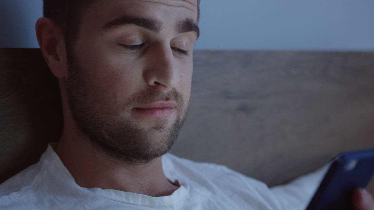 Close-up view of smiling Caucasian man lying in the bed, chatting on the smartphone late at night