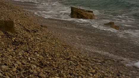 Close-up-of-sea-waves-splashing-on-cliffs-and-pebbles-beach-in-Ionian-coastline