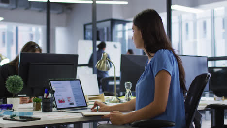 mujer de negocios caucásica sentada en un escritorio usando una computadora portátil y escribiendo notas en un cuaderno en una oficina ocupada