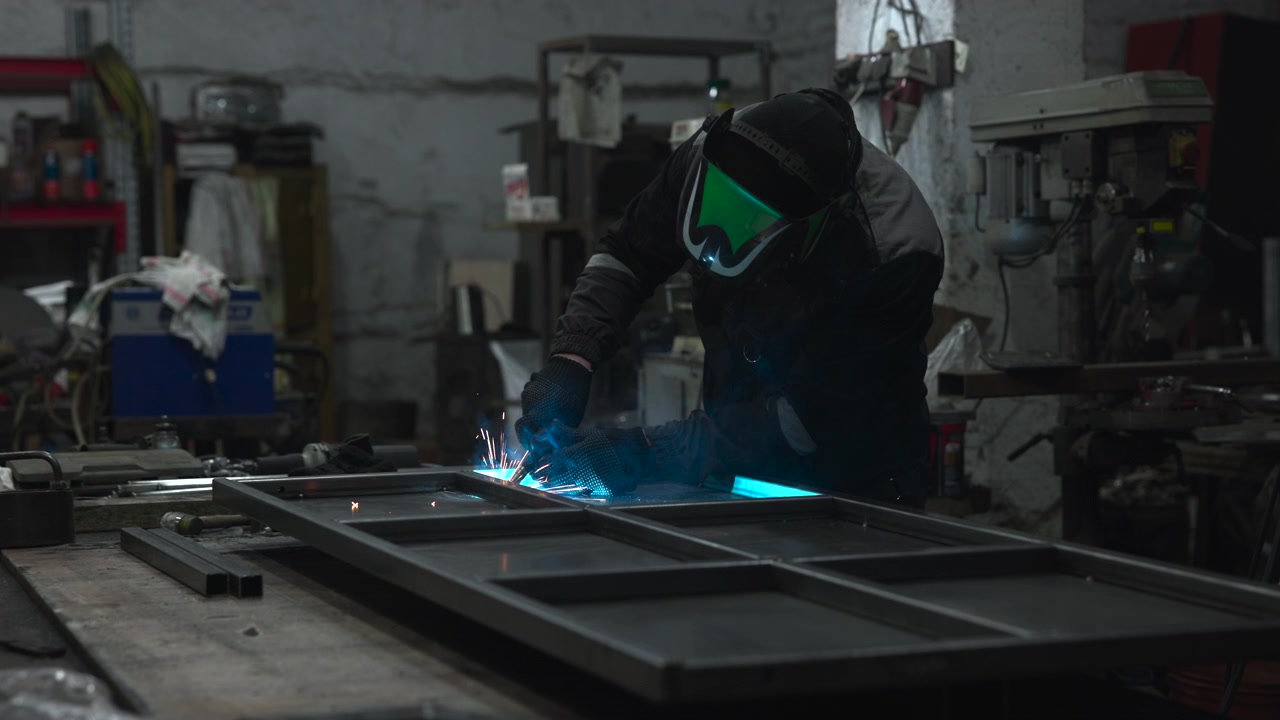 Worker in protective welding helmet and gloves leans over steel structure inside cluttered industrial workshop, with bright blue welding glow and sparks, rough concrete walls, machinery, tools, and