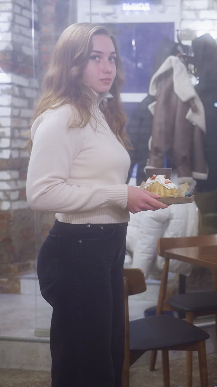 Young girl in beige shirt and black jeans, holding coffee cup and croissant in cafe, sitting close to window. Cozy cafe ambiance with soft lighting and blurred cityscape in background