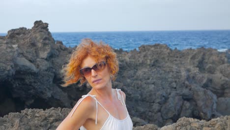 a shot of a redhead woman in white on a cliff beside the ocean, in a volcanic island