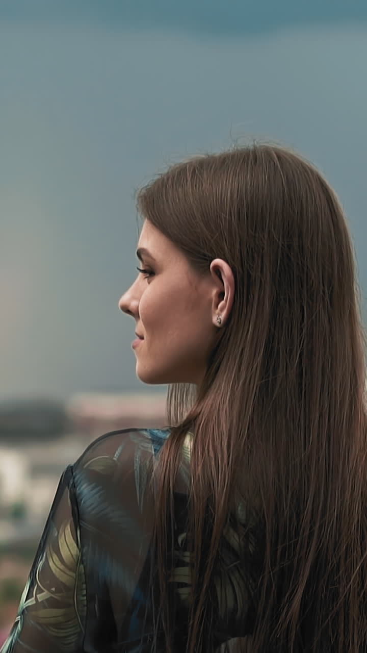 Attractive woman with long loose hair in stylish clothes with color print standing against blurred city on rainy day slow motion close view