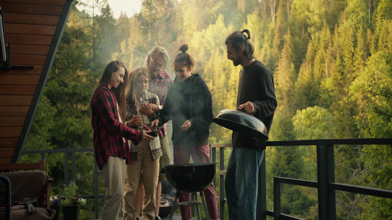 Friends at a picnic: a girl puts sausages on a wood-fired grill, her friends help her in this. Barbecue on the balcony of a country house in the mountains with a coniferous forest
