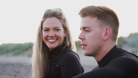 couple wearing wetsuits sitting on beach talking and looking out to sea together